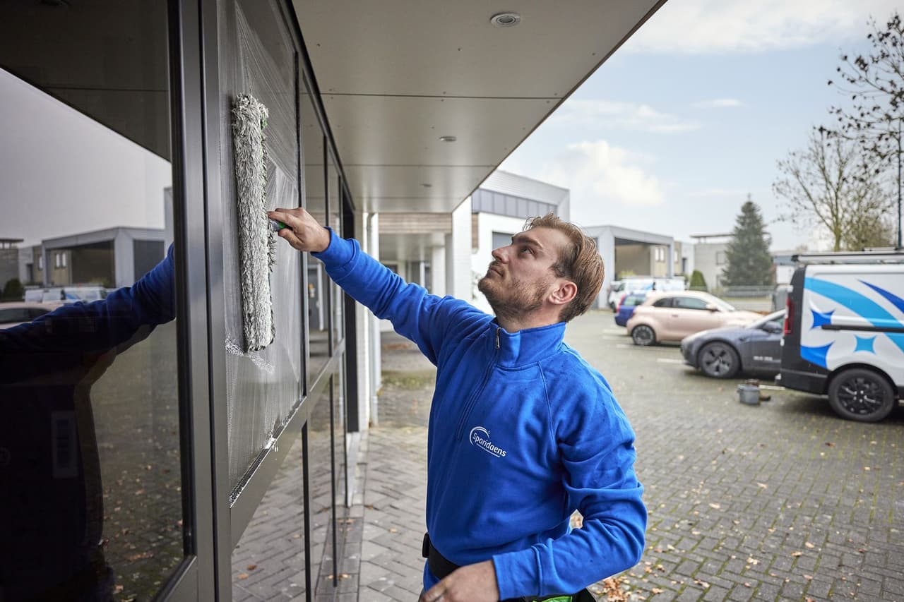 Medewerker van Sparidaens wast de ramen van een bedrijfspand, met de bedrijfsbus op de achtergrond